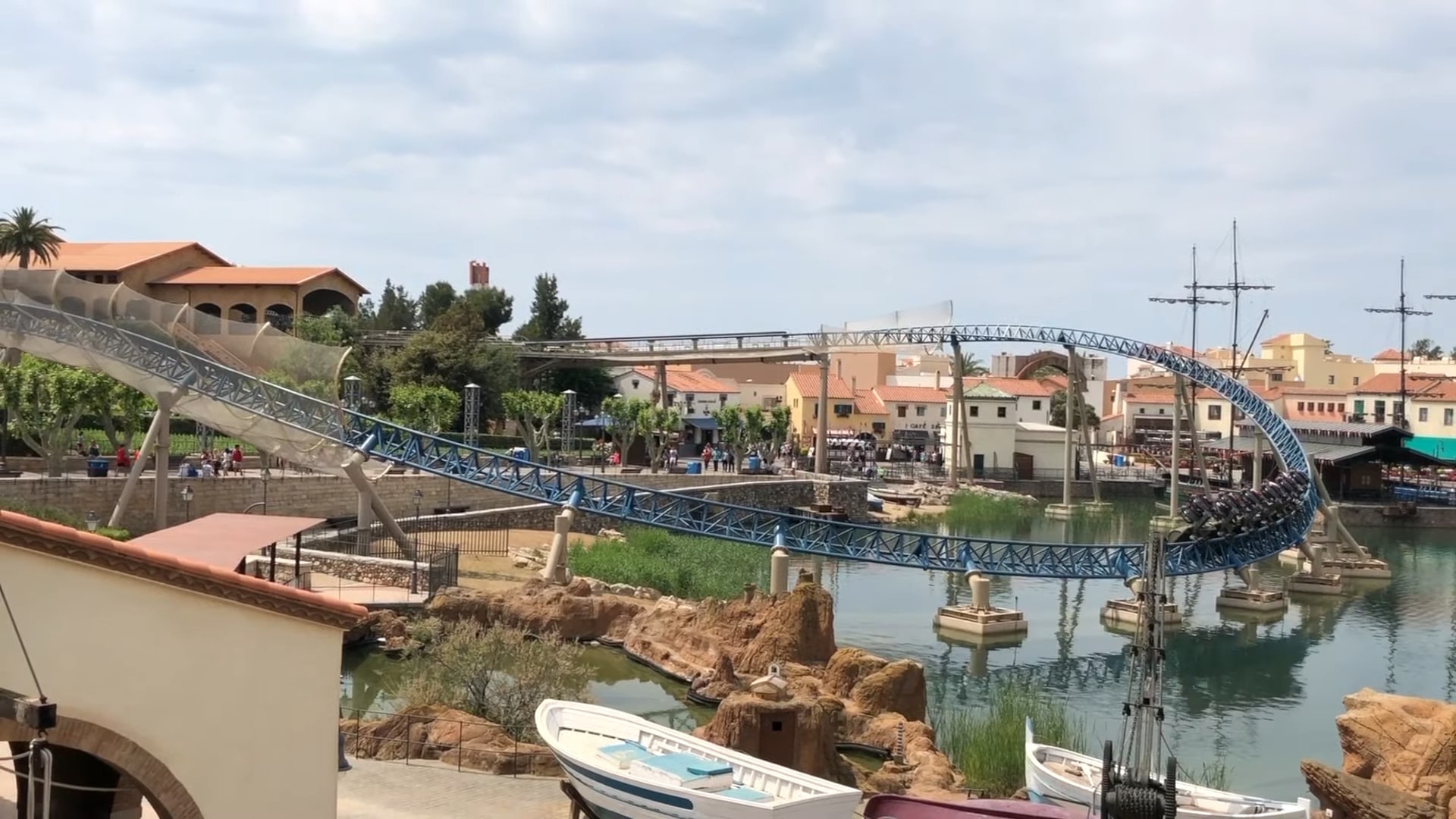 A blue roller coaster with a twisting track over water at PortAventura World in Spain, surrounded by Mediterranean-style buildings